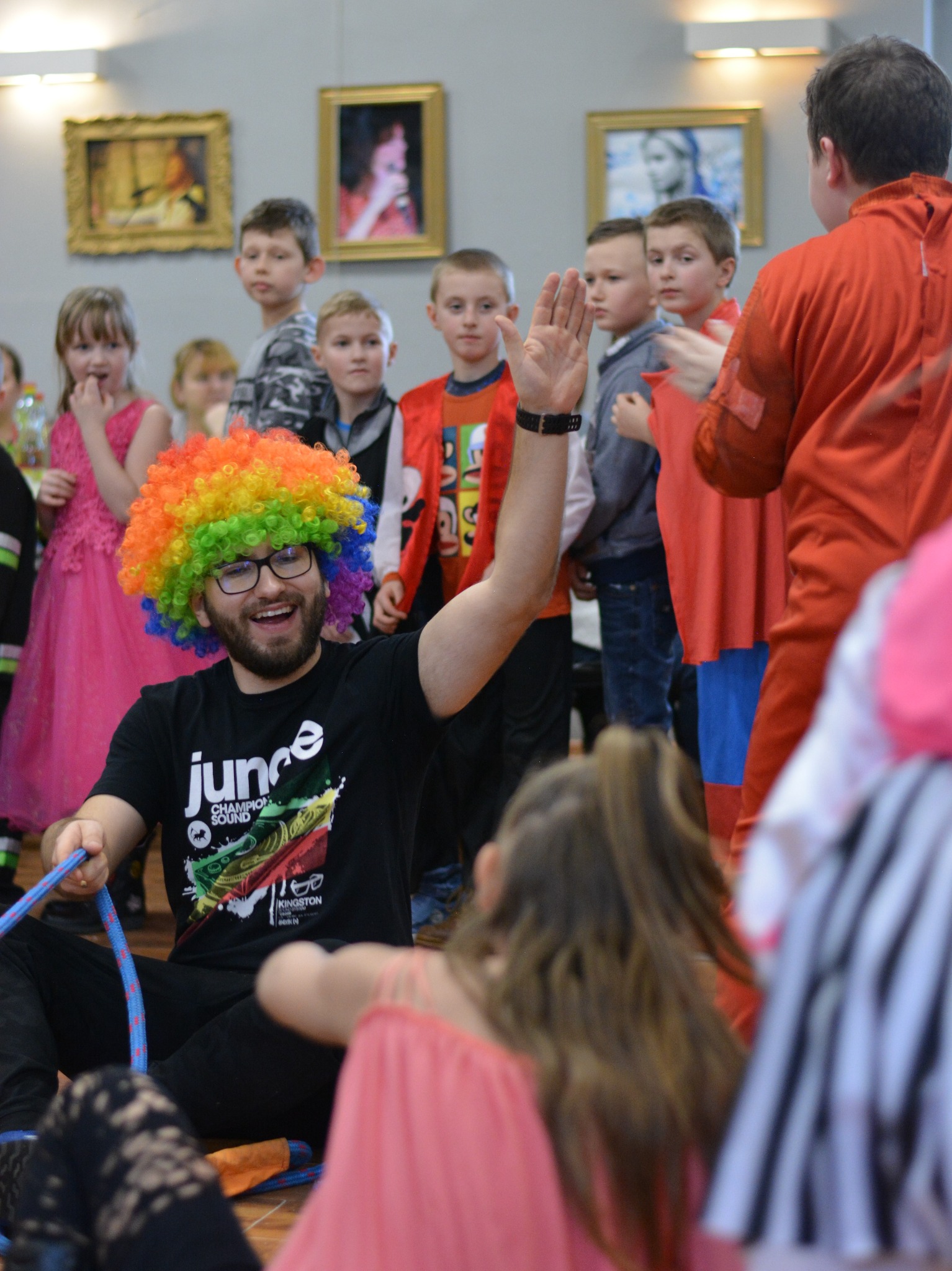Uśmiechnięty mężczyzna w okularach i tęczowej peruce siedzi na podłodze z niebieską liną, machając ręką do grupy dzieci podczas imprezy w pomieszczeniu z obrazami na ścianach.