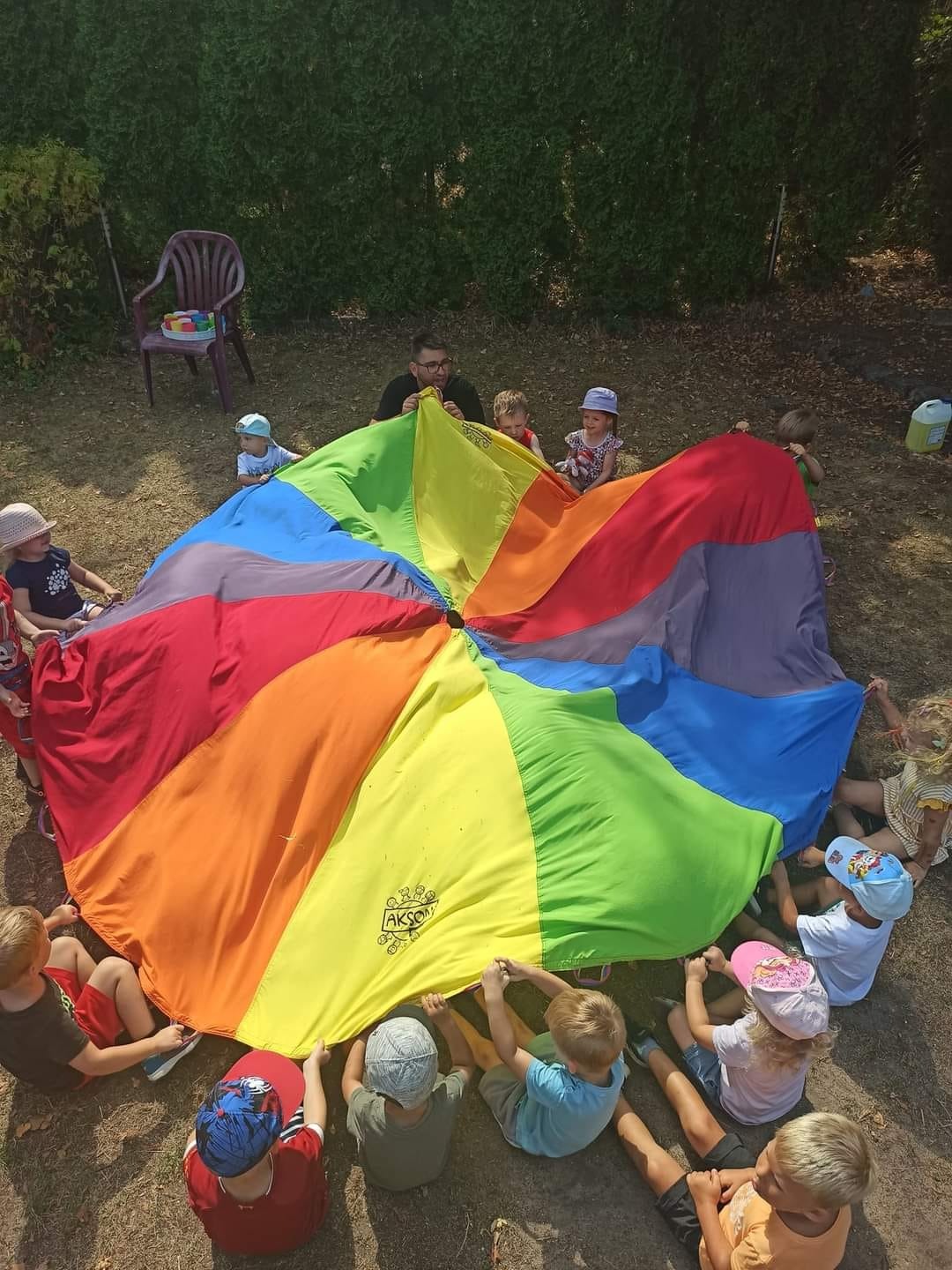 Animator prowadzi zabawę z kolorową chustą animacyjną dla grupy dzieci siedzących na trawie w ogrodzie.