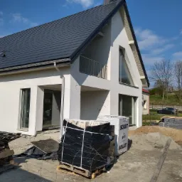 Nowoczesny, biały dom jednorodzinny z ciemnoszarą dachówką, balkonem z metalową balustradą i trójkątnym oknem, w trakcie budowy, z paletami cegieł Solbet na placu budowy i stertą piasku.