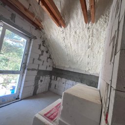 Delta Pur Izolacje - Poddasze ocieplone pianką PUR z widocznymi elementami konstrukcyjnymi dachu i oknem. Ściany z bloczków, częściowo ocieplone. Widoczne materiały budowlane.