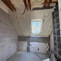Delta Pur Izolacje - Poddasze z oknem połaciowym po natrysku pianki PUR, widoczne drewniane elementy konstrukcji dachu i fragment ściany z bloczków. Biała, porowata struktura pianki.