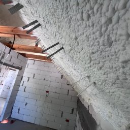 Delta Pur Izolacje - Poddasze ocieplone pianką PUR z widocznymi elementami mocującymi i fragmentami drewnianej konstrukcji. Biała pianka pokrywa skośną powierzchnię.