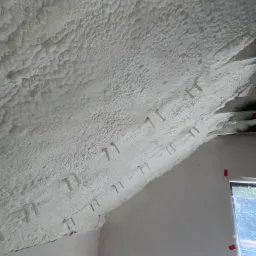 Poddasze ocieplone białą pianką PUR, widoczne mocowania, fragment okna z czerwoną taśmą. Struktura pianki nier&oacute;wna, porowata. Ściana pomalowana na biało.