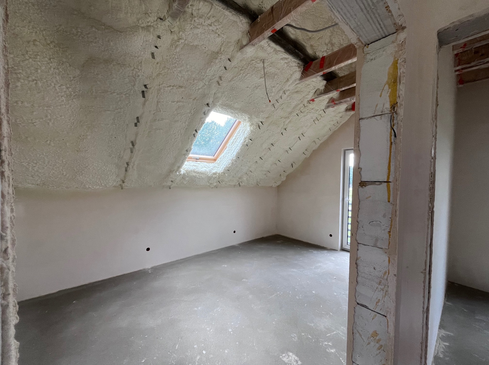 Poddasze po ociepleniu pianką PUR z widocznym oknem dachowym i surowym wykończeniem ścian oraz wylewką podłogową. Widoczne elementy konstrukcji dachu.