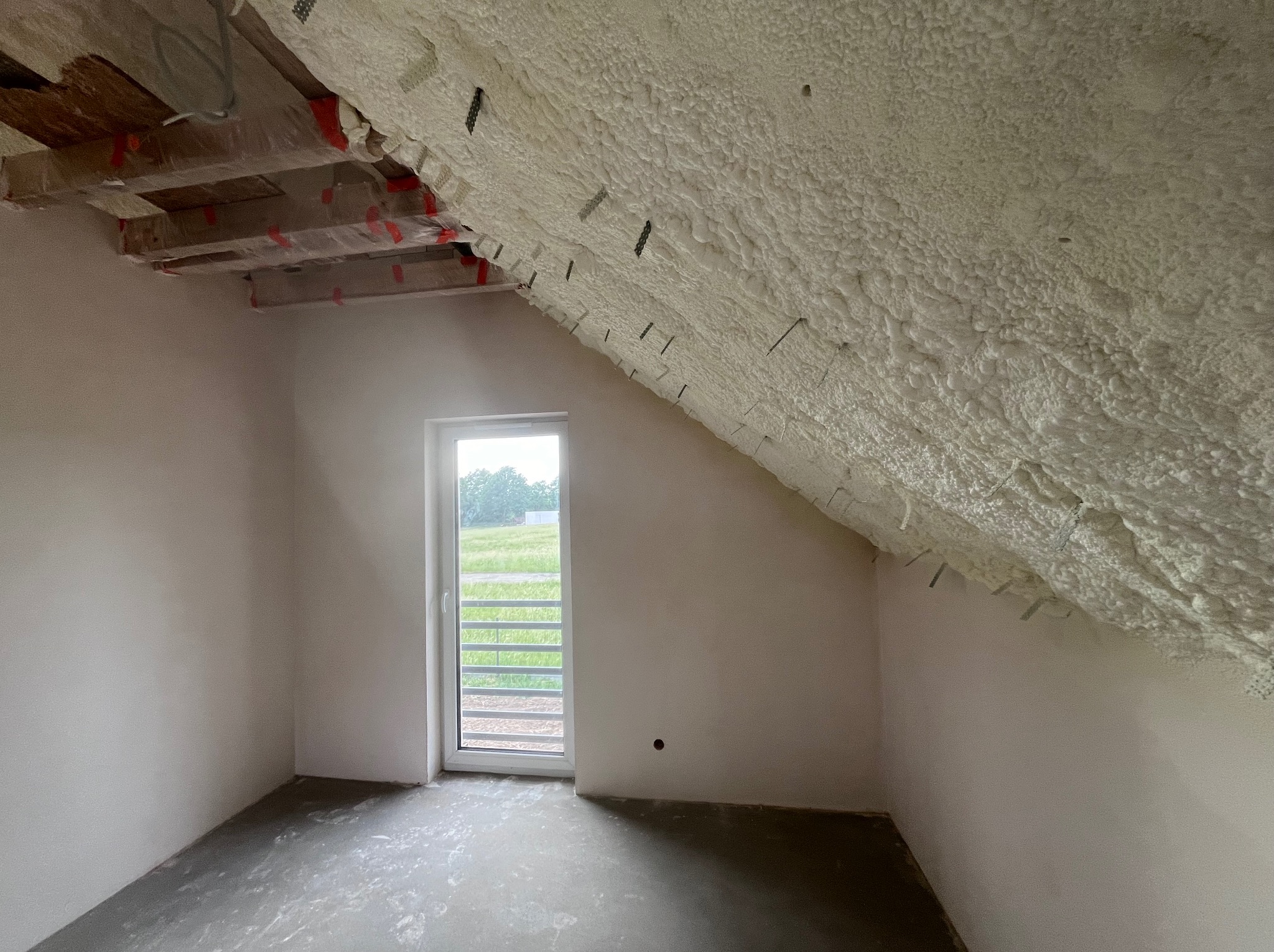 Poddasze po ociepleniu pianką PUR, widoczne krokwie i biała, porowata struktura izolacji. Okno z widokiem na zieleń. Świeżo wylana podłoga.