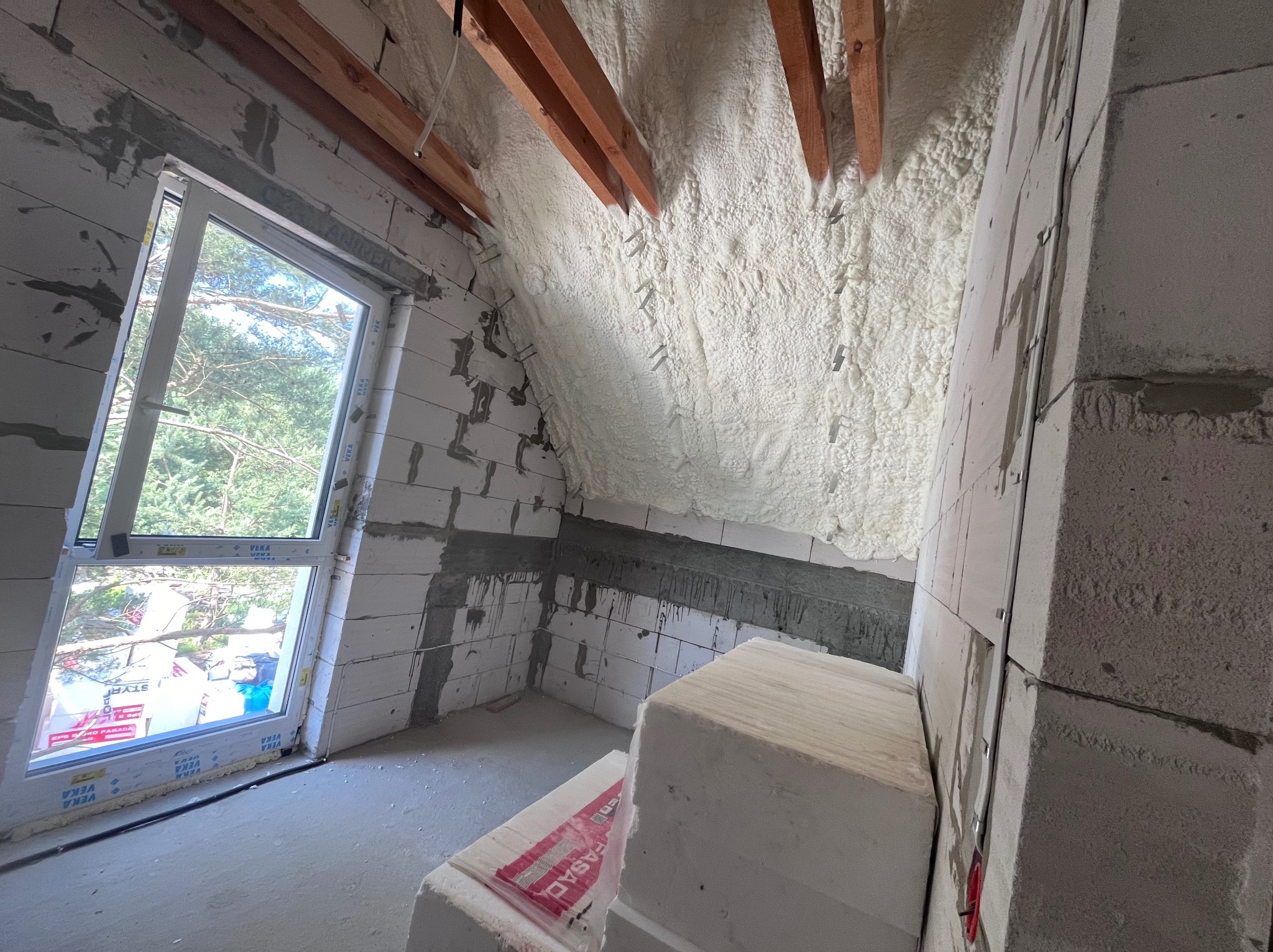 Poddasze ocieplone pianką PUR z widocznymi elementami konstrukcyjnymi dachu i oknem. Ściany z bloczków, częściowo ocieplone. Widoczne materiały budowlane.