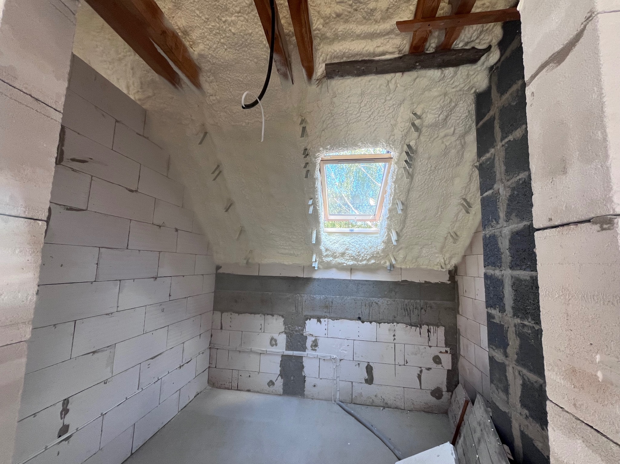 Poddasze z oknem połaciowym po natrysku pianki PUR, widoczne drewniane elementy konstrukcji dachu i fragment ściany z bloczków. Biała, porowata struktura pianki.