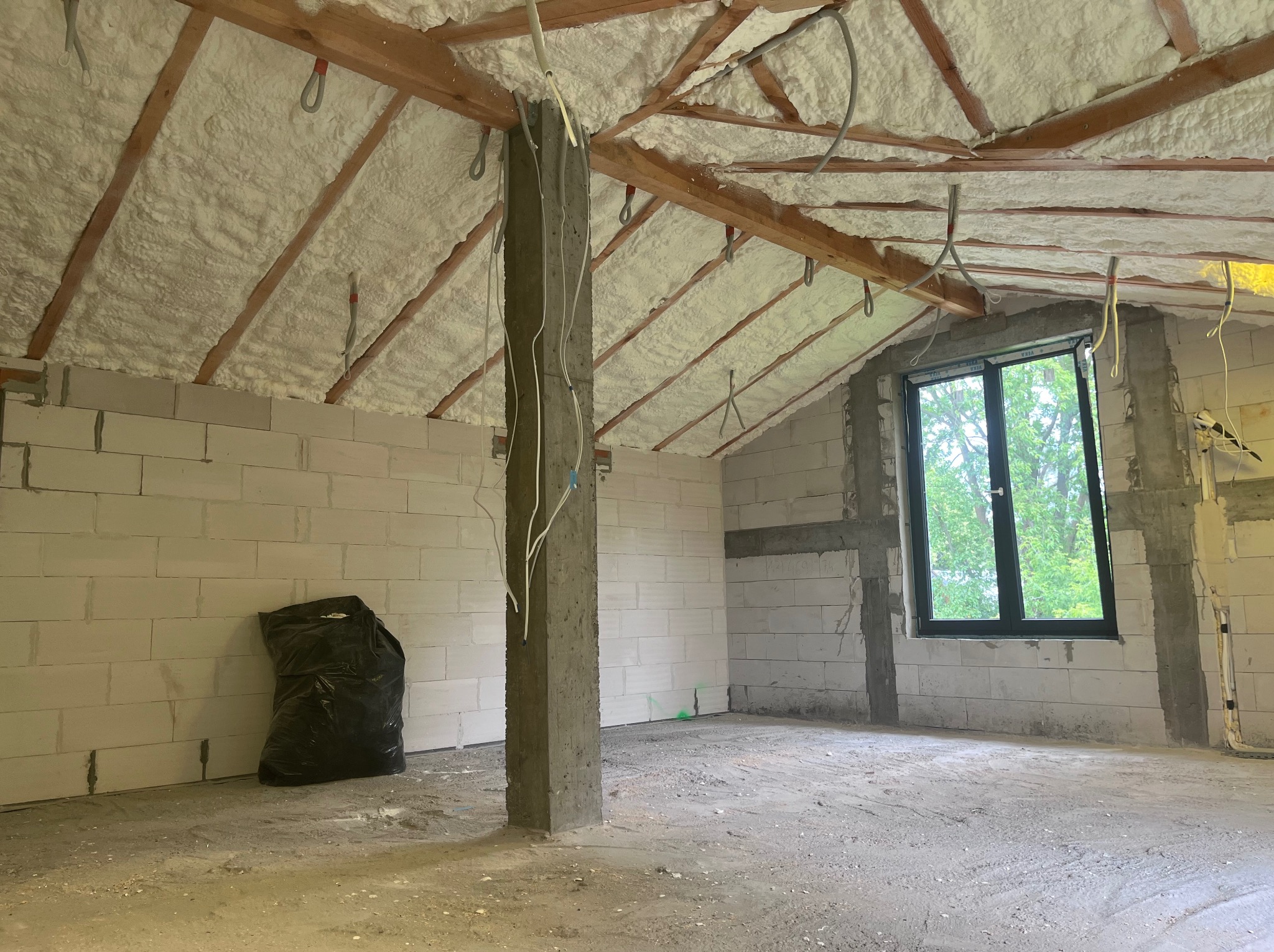 Poddasze w trakcie ocieplania pianką PUR. Widoczne białe bloki, okno i czarny worek na podłodze. Na krokwiach drewnianych naniesiona pianka.