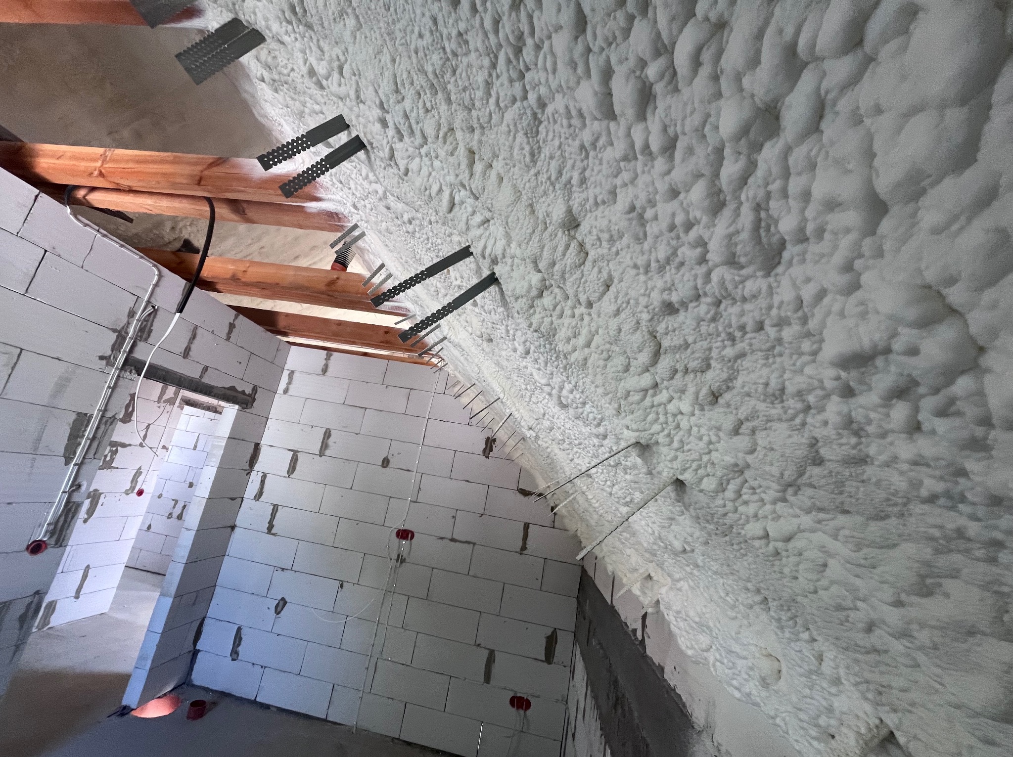Poddasze ocieplone pianką PUR z widocznymi elementami mocującymi i fragmentami drewnianej konstrukcji. Biała pianka pokrywa skośną powierzchnię.