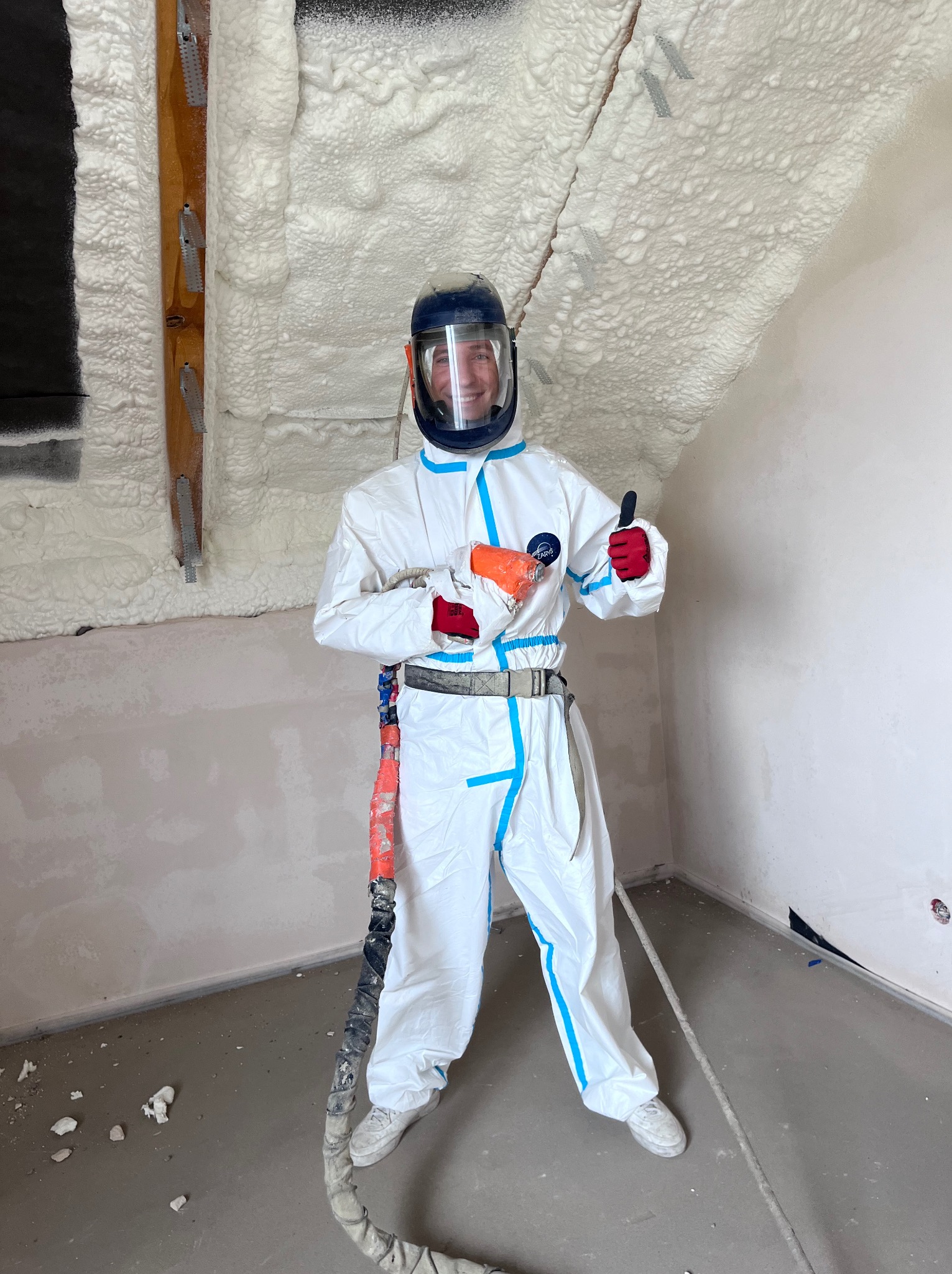 Pracownik w kombinezonie po ociepleniu poddasza pianką PUR w Nadmie, z pistoletem natryskowym i kciukiem w górę. Ściany pokryte pianą.
