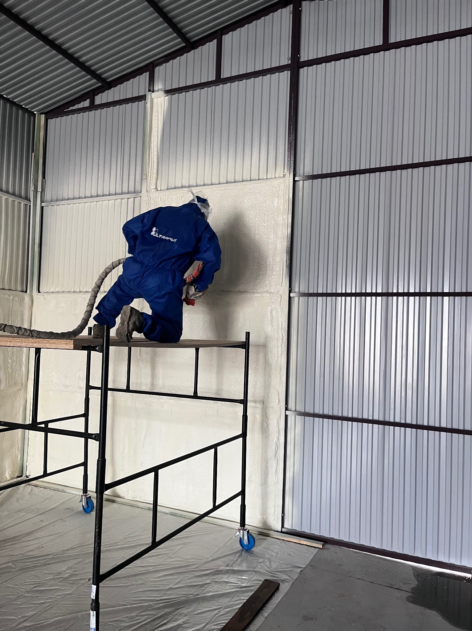 Pracownik w kombinezonie aplikuje piankę PUR na metalową konstrukcję, stojąc na rusztowaniu. Widoczne świeżo nałożone ocieplenie i narzędzie do natrysku.