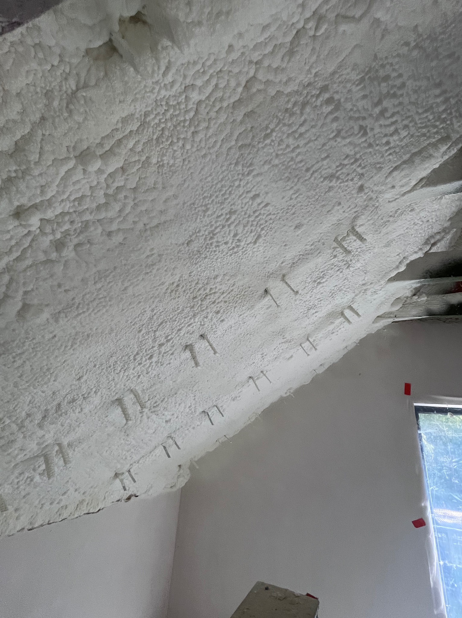 Poddasze ocieplone białą pianką PUR, widoczne mocowania, fragment okna z czerwoną taśmą. Struktura pianki nierówna, porowata. Ściana pomalowana na biało.