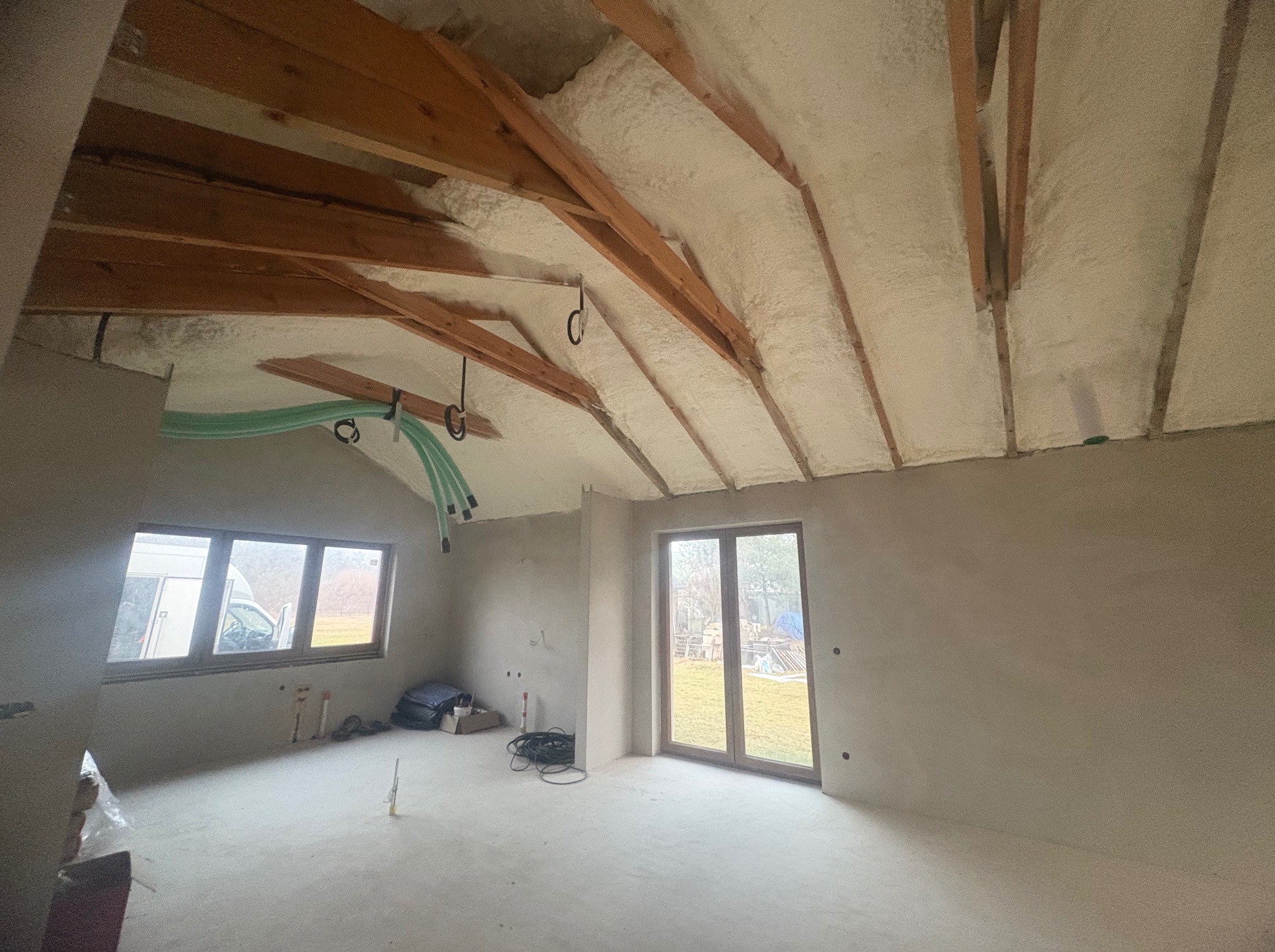 Poddasze po ociepleniu pianką PUR: widoczne drewniane belki stropowe i szara pianka. Na ścianach widoczne instalacje. Dwa okna z widokiem na zewnątrz.