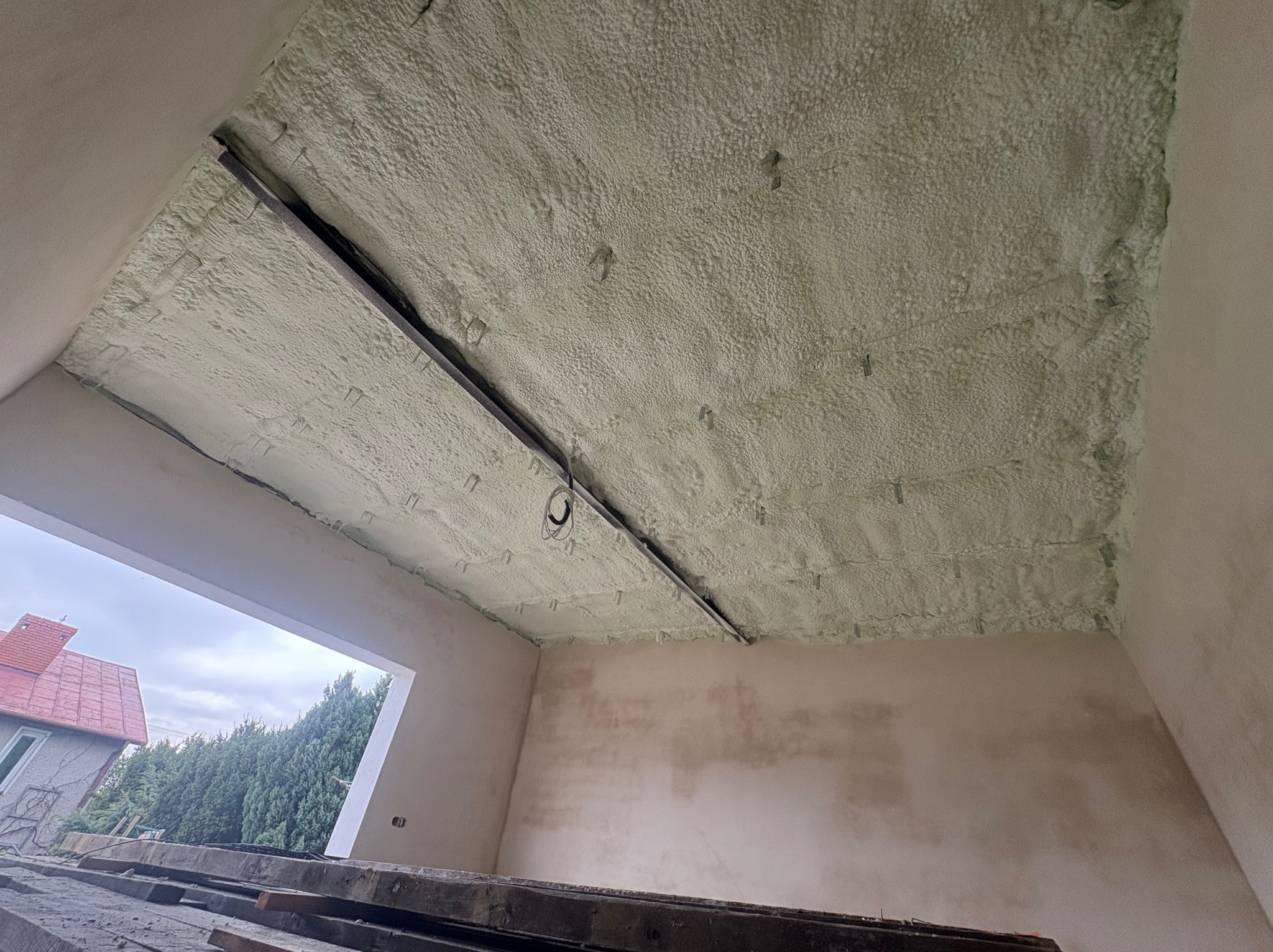 Poddasze po ociepleniu pianką PUR, widoczne elementy konstrukcyjne i wystająca instalacja. Na zewnątrz budynek z czerwoną dachówką i zieleń.