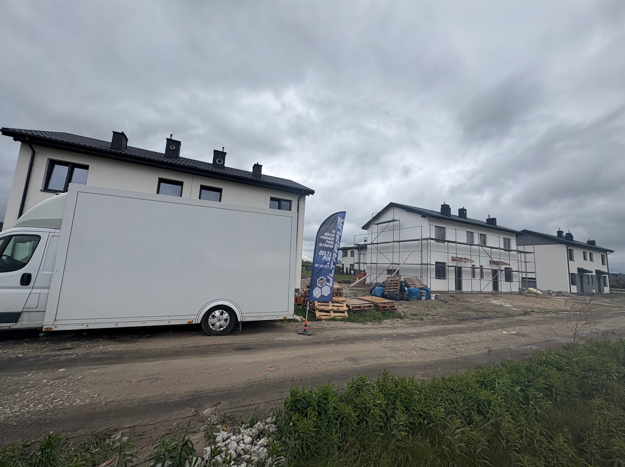Ocieplone budynki w zabudowie szeregowej, widoczne rusztowania i samochód dostawczy z banerem reklamowym pianki PUR. Pochmurne niebo.