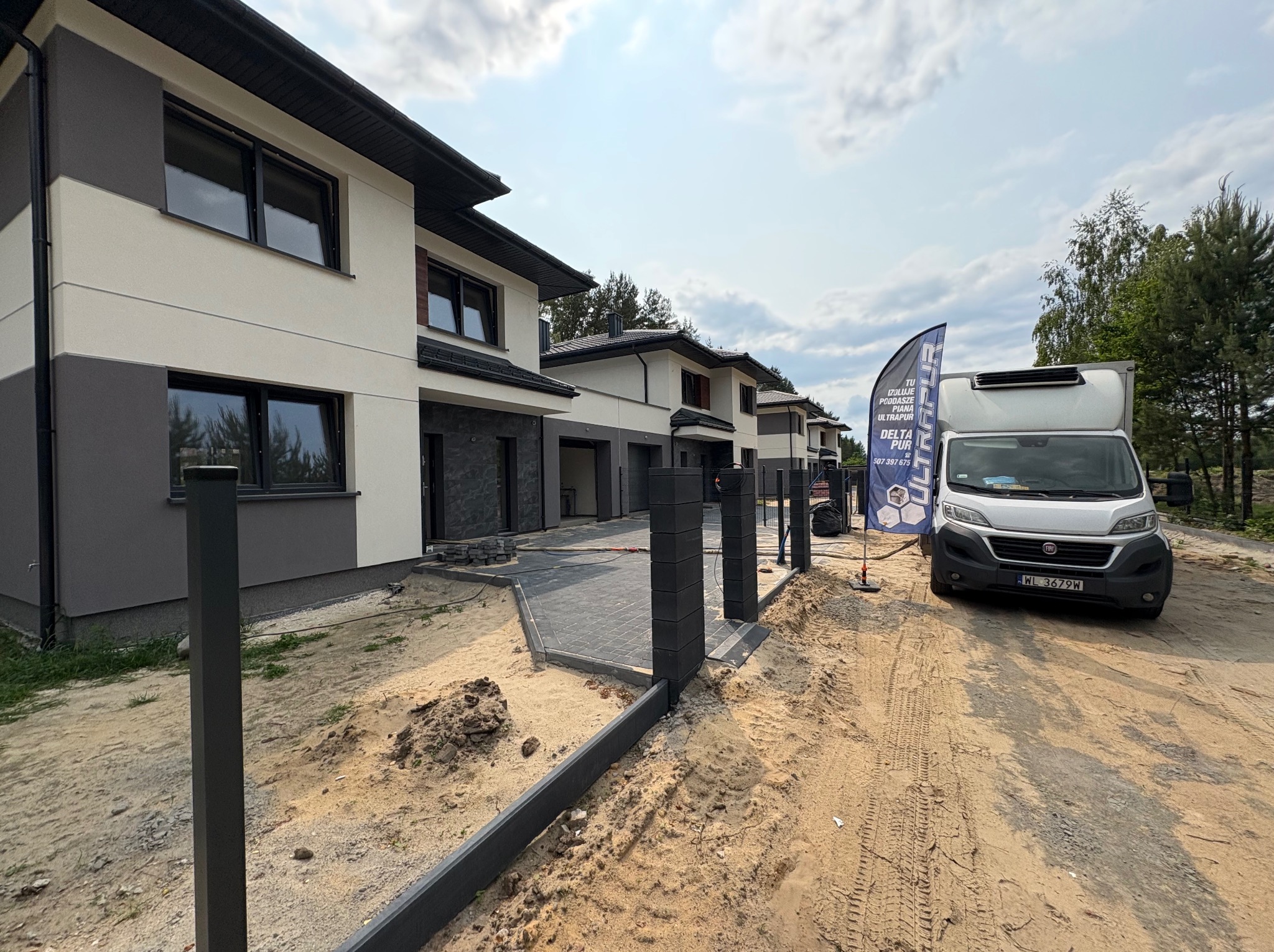 Nowoczesne domy w zabudowie szeregowej z szarą elewacją i czarną stolarką okienną. Na pierwszym planie utwardzona kostką ścieżka i firmowy van Delta Pur.