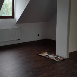 Poddasze z ułożonymi ciemnymi panelami podłogowymi, widoczne fragmenty paneli na kartonie, grzejnik pod oknem i surowy otwór drzwiowy.