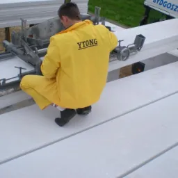 Pracownik w żółtym kombinezonie z napisem 'YTONG' na plecach, kuca przy białych bloczkach z betonu komórkowego, używając specjalistycznego urządzenia do ich przenoszenia i układania.