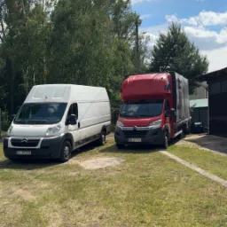 Dwa busy transportowe: biały furgon i czerwony z plandeką, stoją na trawniku przed budynkiem w słoneczny dzień. Widok na transport i przeprowadzkę.