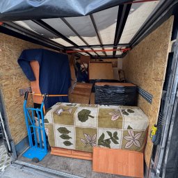 Kacper Kacper - Wnętrze przestrzeni ładunkowej busa wypełnione meblami i przedmiotami, zabezpieczonymi do transportu podczas przeprowadzki. Widoczny wózek transportowy.