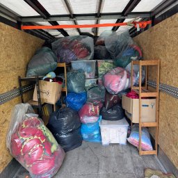 Kacper Kacper - Wnętrze busa wypełnione po brzegi spakowanymi rzeczami w workach i kartonach, zabezpieczonymi pasami transportowymi. Widoczne półki z zapakowanymi przedmiotami.