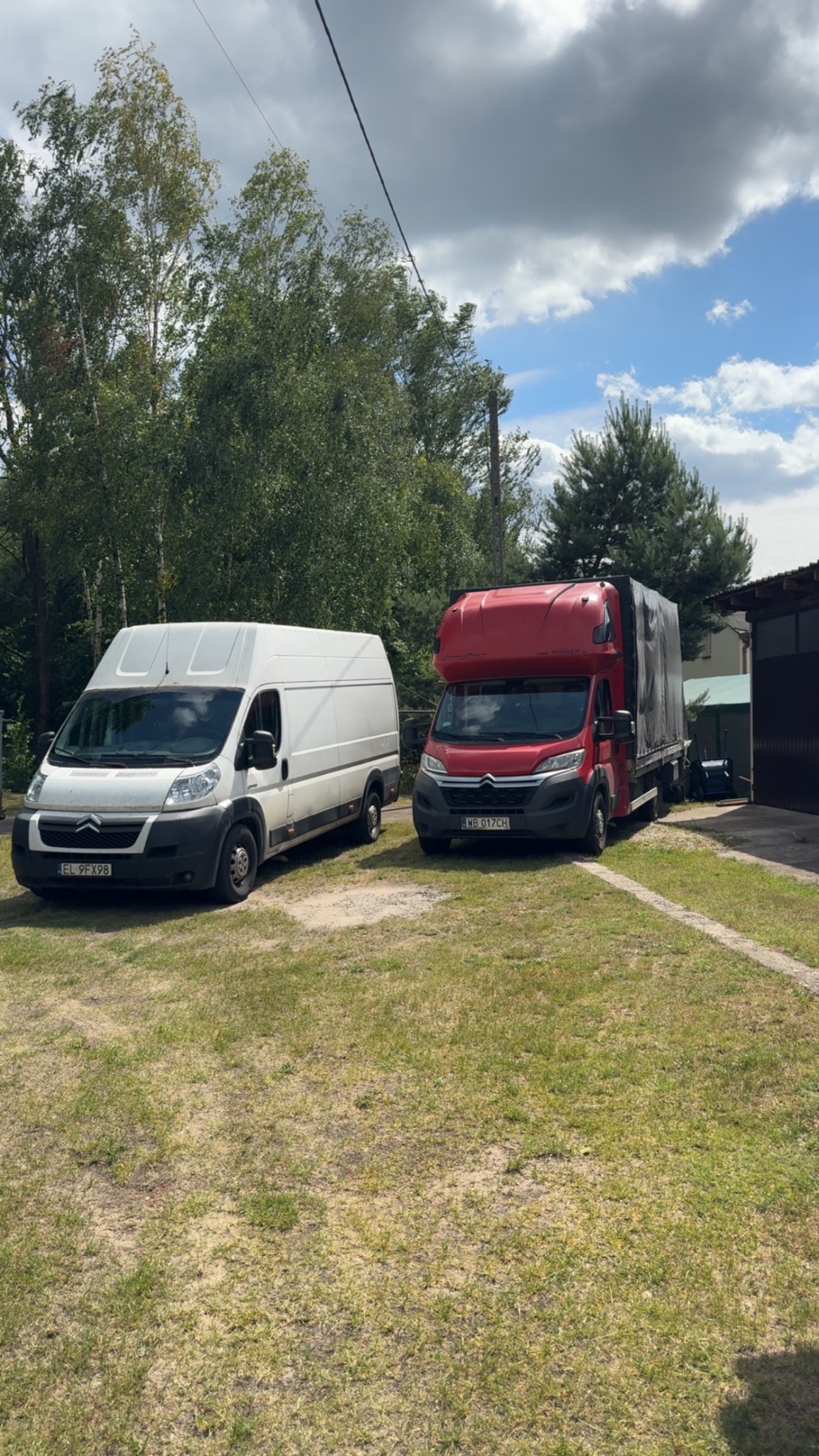 Dwa busy transportowe: biały furgon i czerwony z plandeką, stoją na trawniku przed budynkiem w słoneczny dzień. Widok na transport i przeprowadzkę.