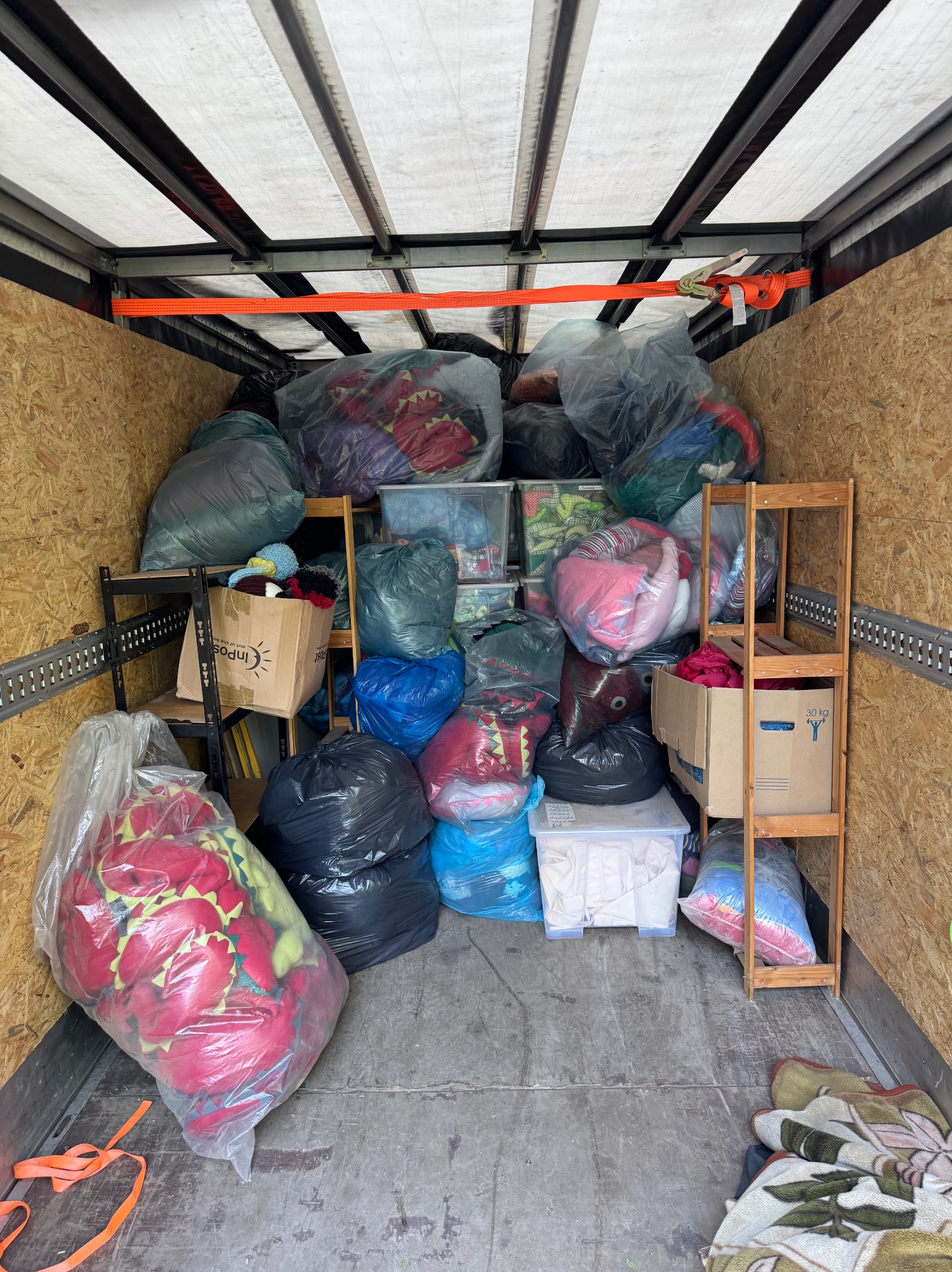 Wnętrze busa wypełnione po brzegi spakowanymi rzeczami w workach i kartonach, zabezpieczonymi pasami transportowymi. Widoczne półki z zapakowanymi przedmiotami.