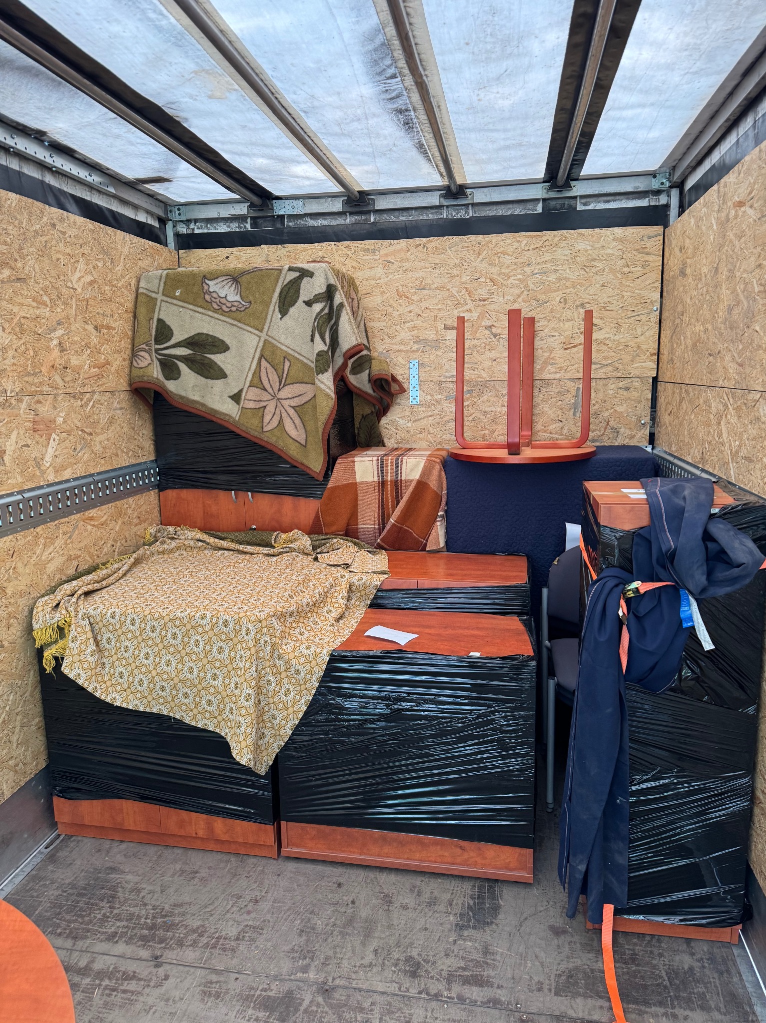 Meble zabezpieczone folią i kocami w przestrzeni ładunkowej busa. Widoczne krzesła i elementy garderoby. Transportowany dobytek przygotowany do przewozu.