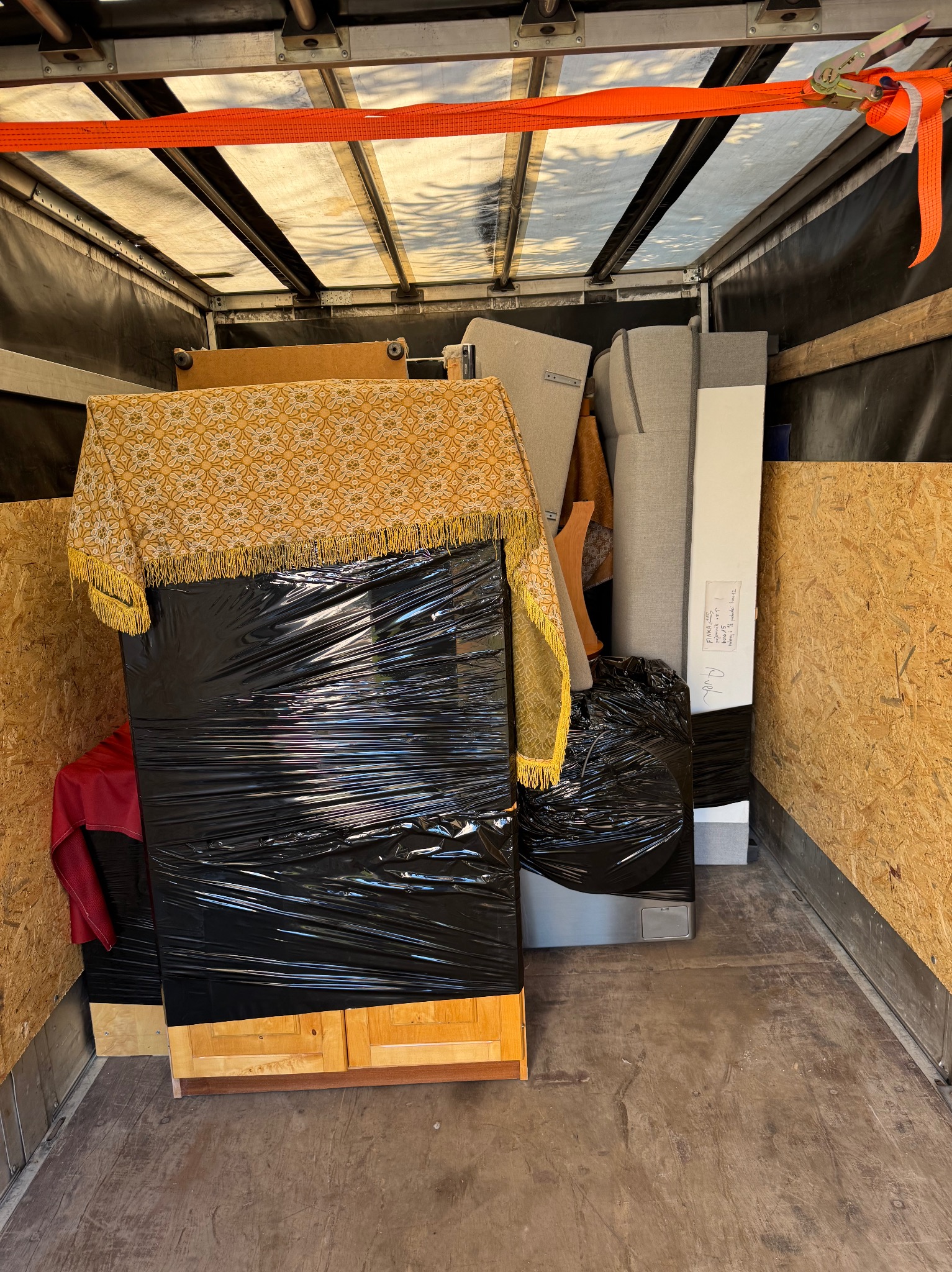 Meble zabezpieczone folią i pasami w przestrzeni ładunkowej busa, gotowe do transportu. Widoczne elementy wyposażenia wnętrz i skrzynia z żółtą narzutą.