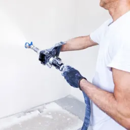 Pracownik w rękawicach natryskowo aplikuje substancję na ścianę za pomocą pistoletu malarskiego; widoczne drobne niedoskonałości podłoża