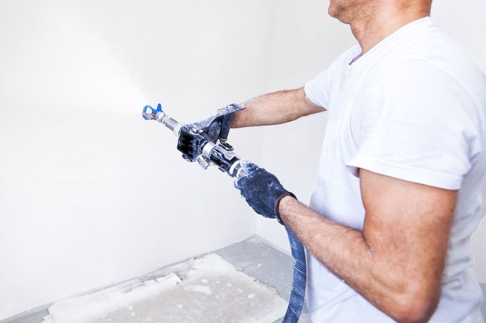 Pracownik w rękawicach natryskowo aplikuje substancję na ścianę za pomocą pistoletu malarskiego; widoczne drobne niedoskonałości podłoża