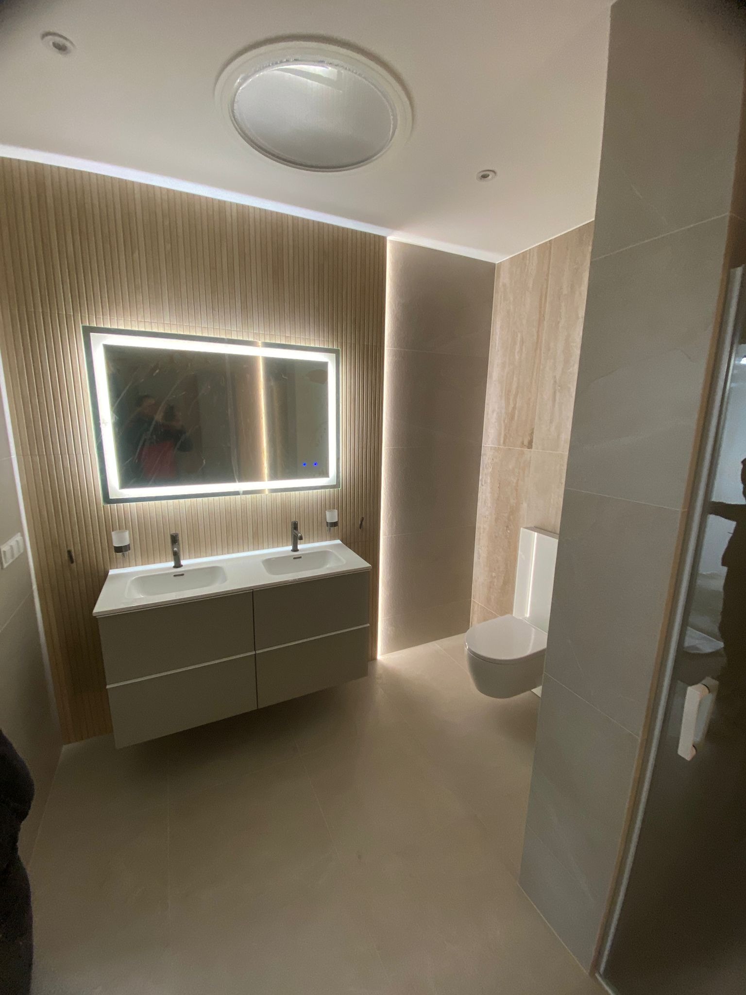 Nowoczesna łazienka z podwójną umywalką na szafce, podświetlanym lustrem, drewnianą ścianą i beżowymi płytkami, oświetleniem sufitowym i toaletą w neutralnej kolorystyce.