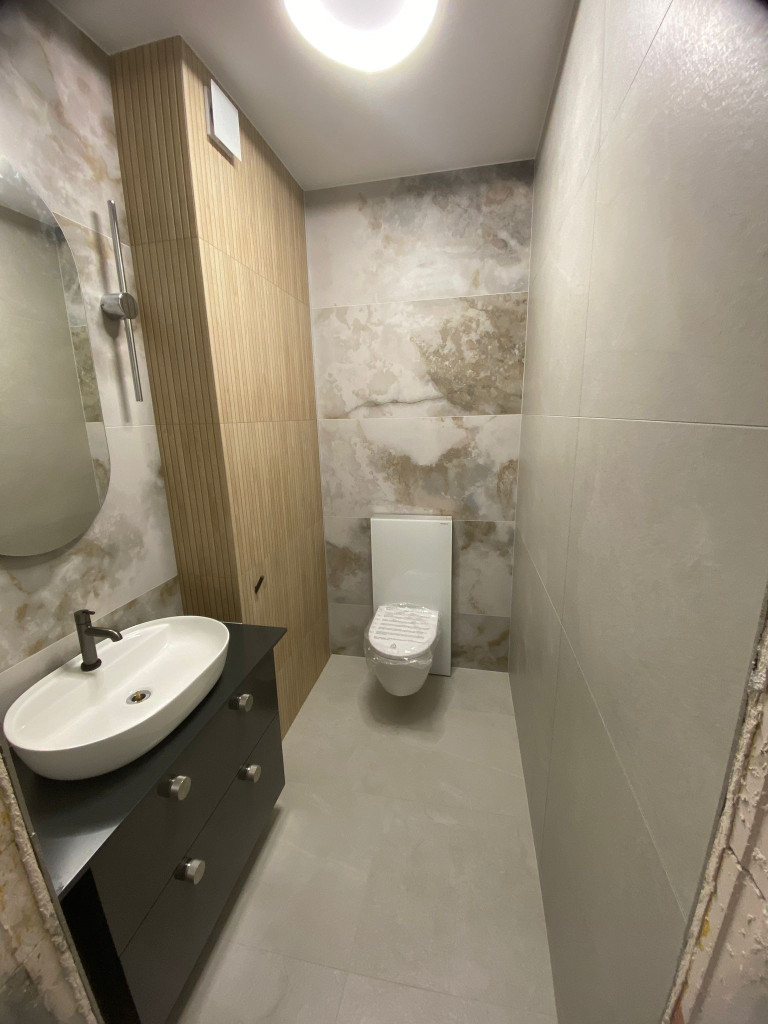 Nowoczesna łazienka z umywalką nablatową na czarnej szafce, toaletą w folii ochronnej i ścianami wykończonymi płytkami w odcieniach beżu i szarości, z elementami dekoracyjnymi z drewnianych paneli.