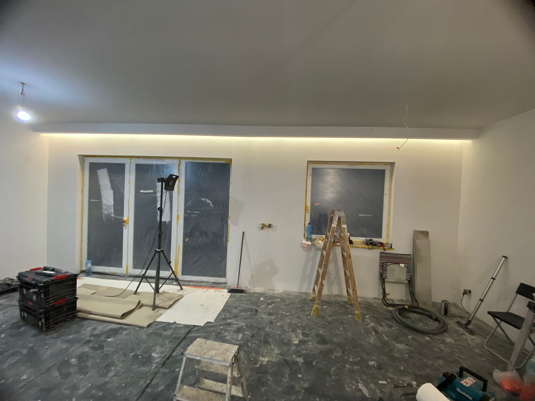 Wnętrze w trakcie prac wykończeniowych: okna i drzwi zabezpieczone folią, drabina, narzędzia, podłoga częściowo zakryta, listwa LED pod sufitem.