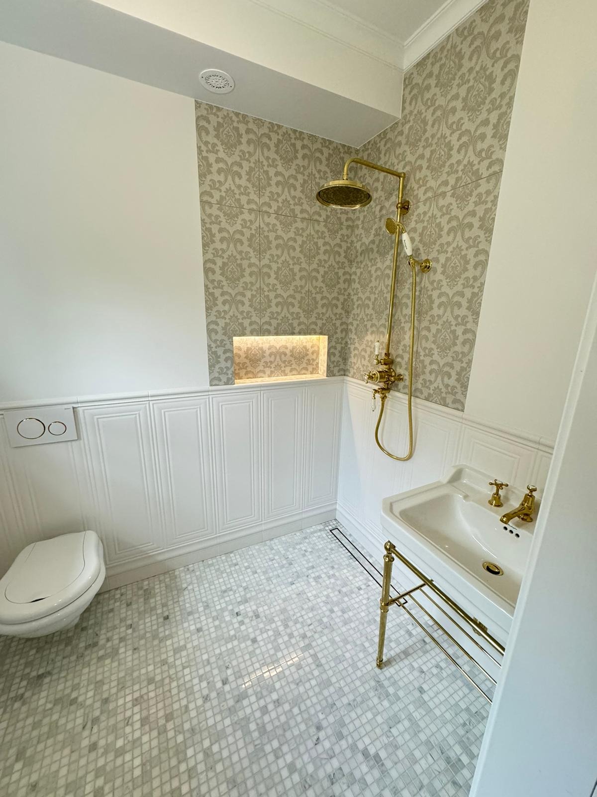 Elegancka łazienka z mozaikową podłogą, białą ceramiką, złotą armaturą i dekoracyjną ścianą z wzorzystymi płytkami.