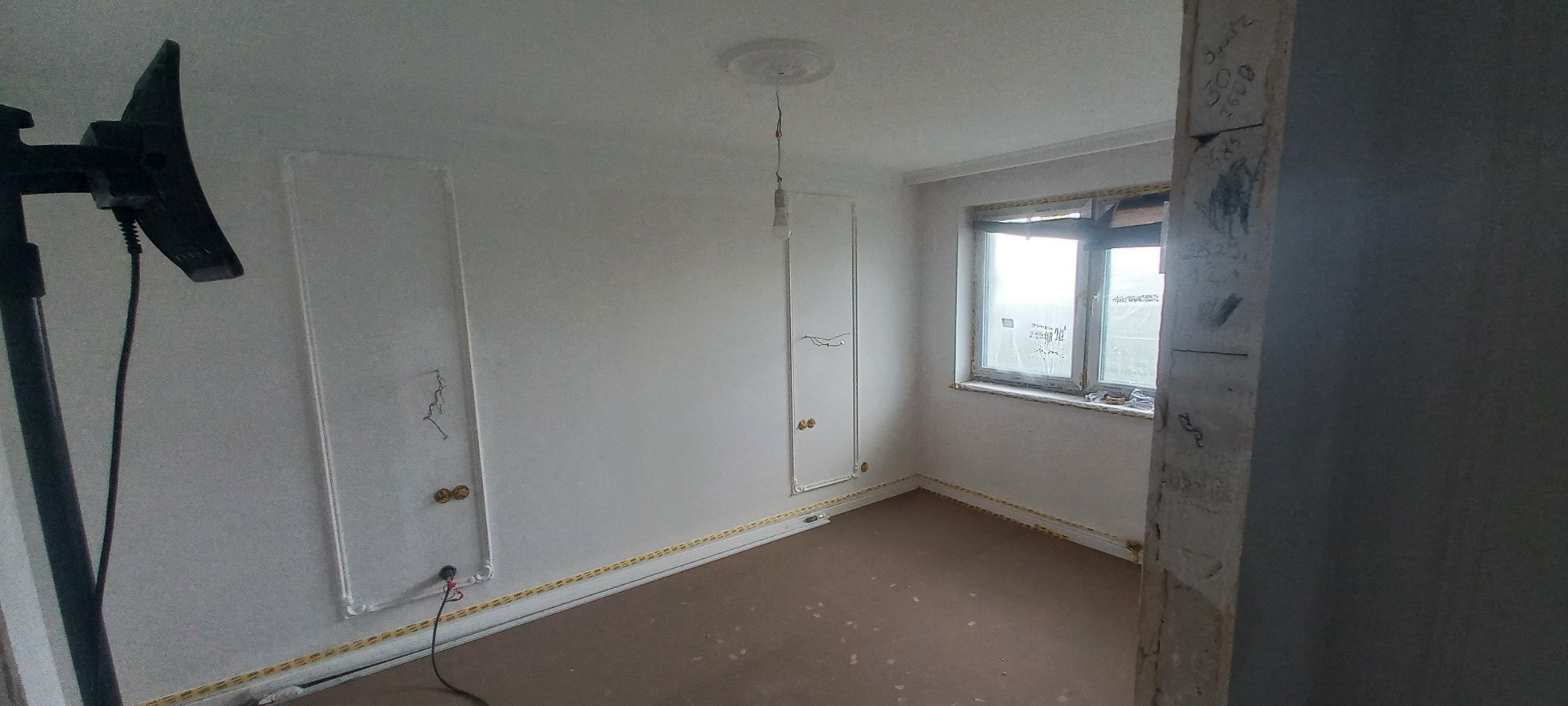 Pokój w trakcie remontu z widocznymi śladami po instalacjach elektrycznych, zabezpieczonymi taśmą przypodłogowymi i oknami, oraz lampą studyjną z lewej strony.