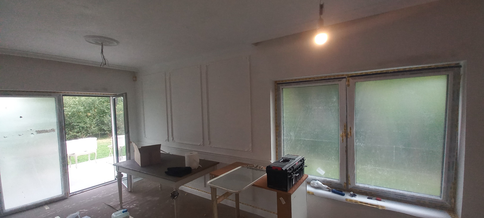 Wnętrze pokoju w trakcie remontu: ściany z dekoracyjnymi listwami, otwarte drzwi balkonowe, okno z widokiem na zieleń, narzędzia i materiały budowlane na stole i parapecie.