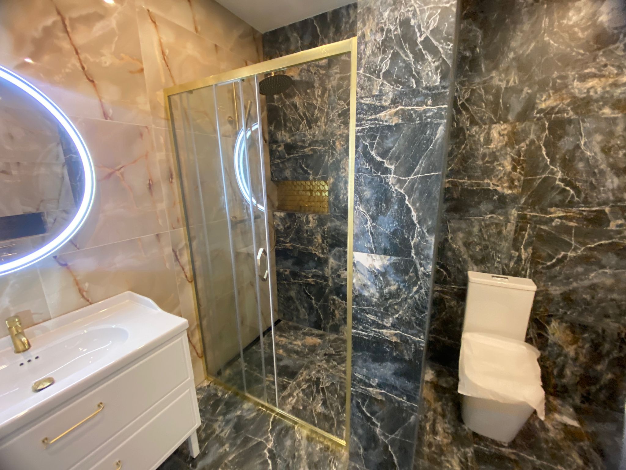 Wykończona łazienka z kabiną prysznicową ze złotymi ramami, umywalką z szafką w stylu retro i toaletą z zabezpieczeniem, ściany wyłożone marmurem w odcieniach brązu i szarości.