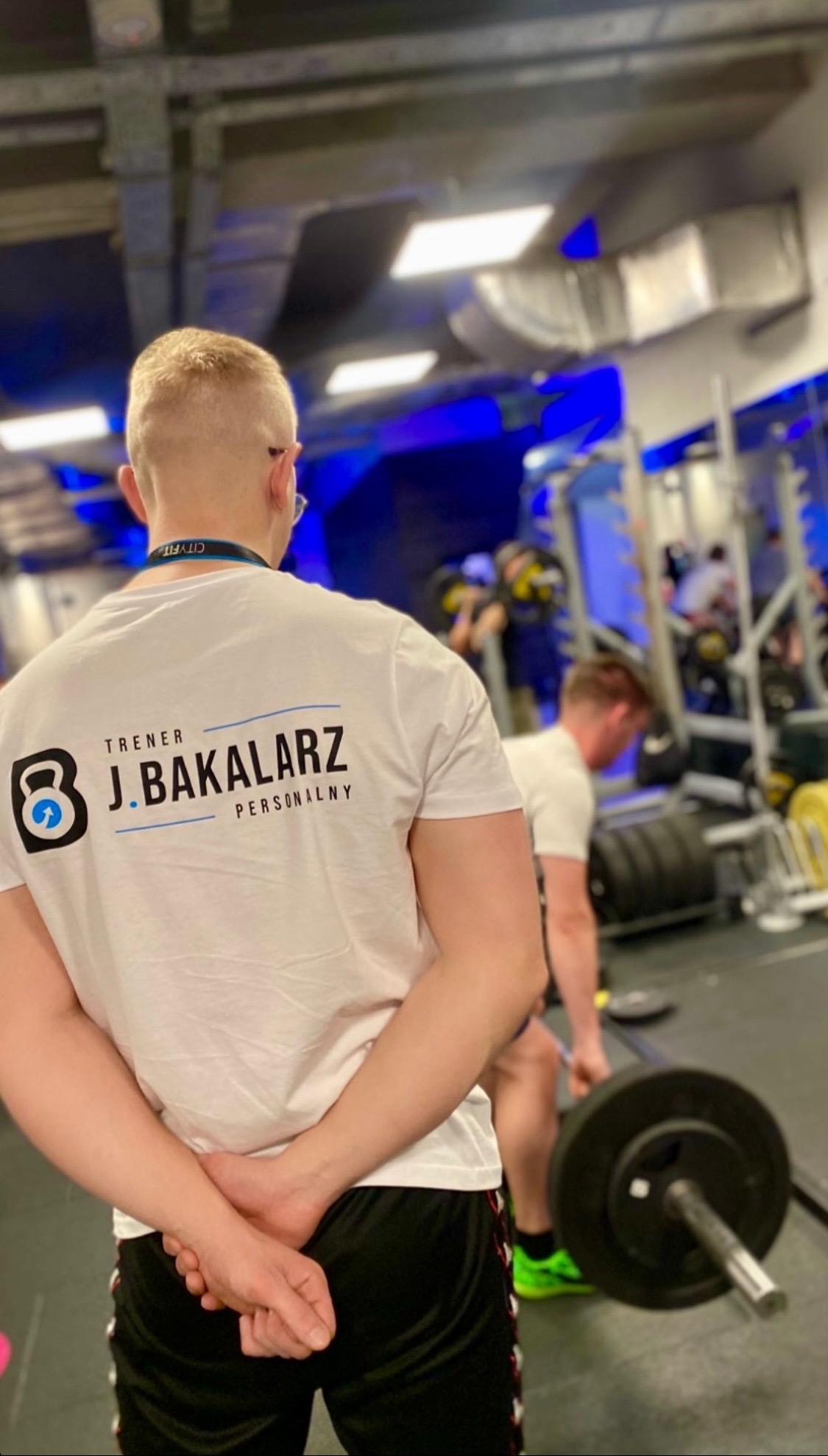 Trener personalny J. Bakalarz z tyłu, w tle osoba podnosząca ciężary na siłowni, widoczny sprzęt do ćwiczeń.