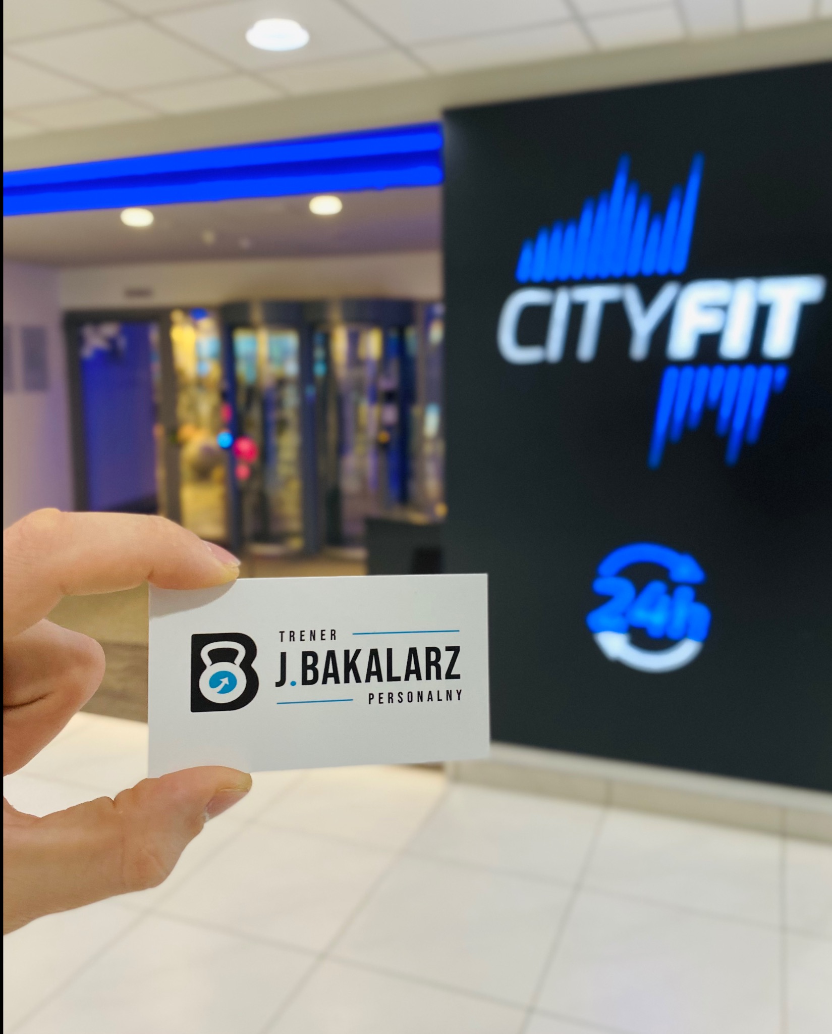 Wnętrze CityFit, w dłoni wizytówka trenera personalnego J. Bakalarz z logo kettlebell i strzałką. W tle wejście do klubu.