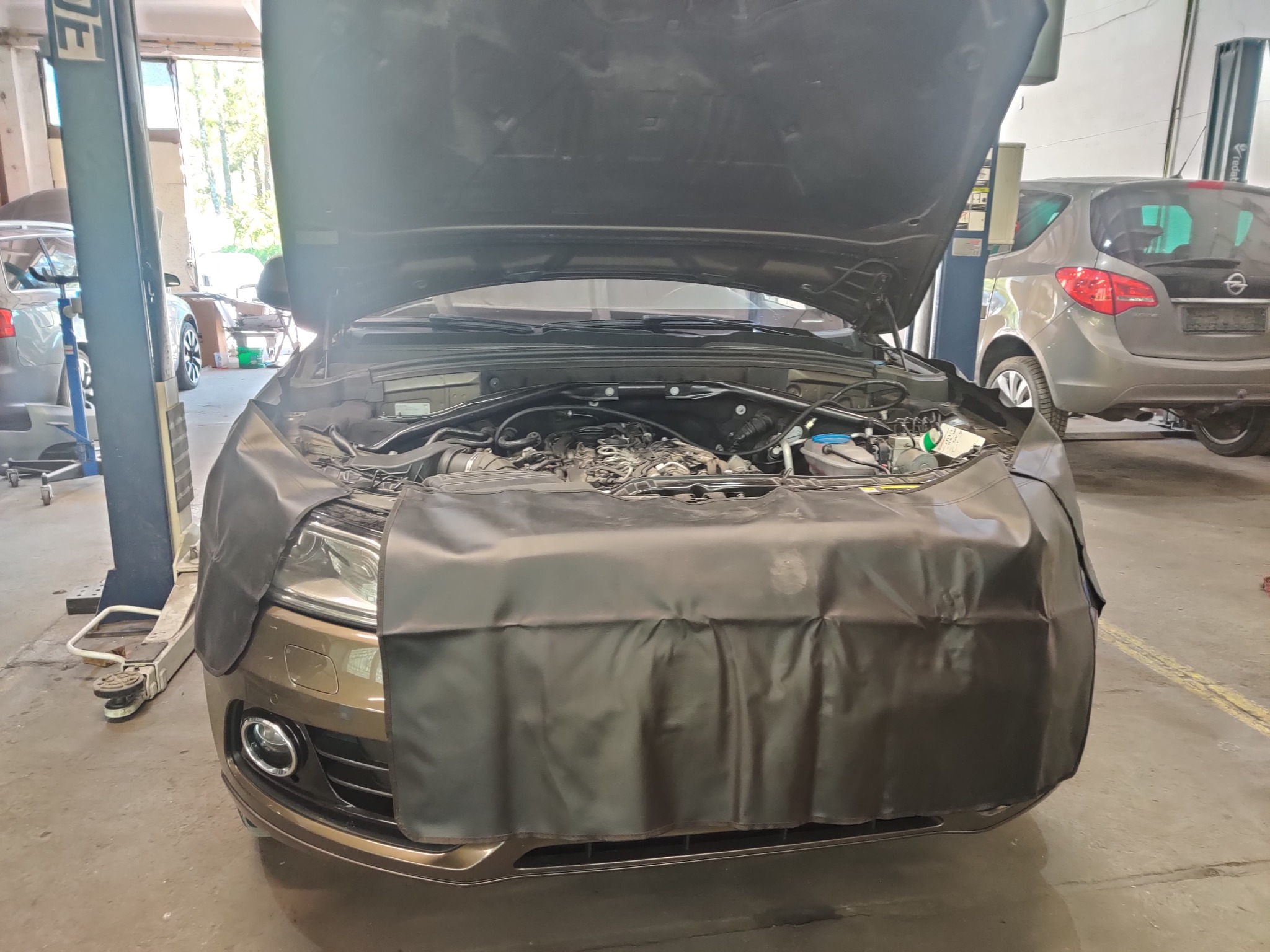Brązowe Audi Q5 z otwartą maską silnika, zabezpieczone czarną osłoną warsztatową podczas prac w warsztacie samochodowym.