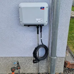 Szara ładowarka EV marki SMA zamontowana na elewacji budynku, z widocznym kablem i złączem, obok rury spustowej i instalacji wodnej na tle drobnego żwiru.