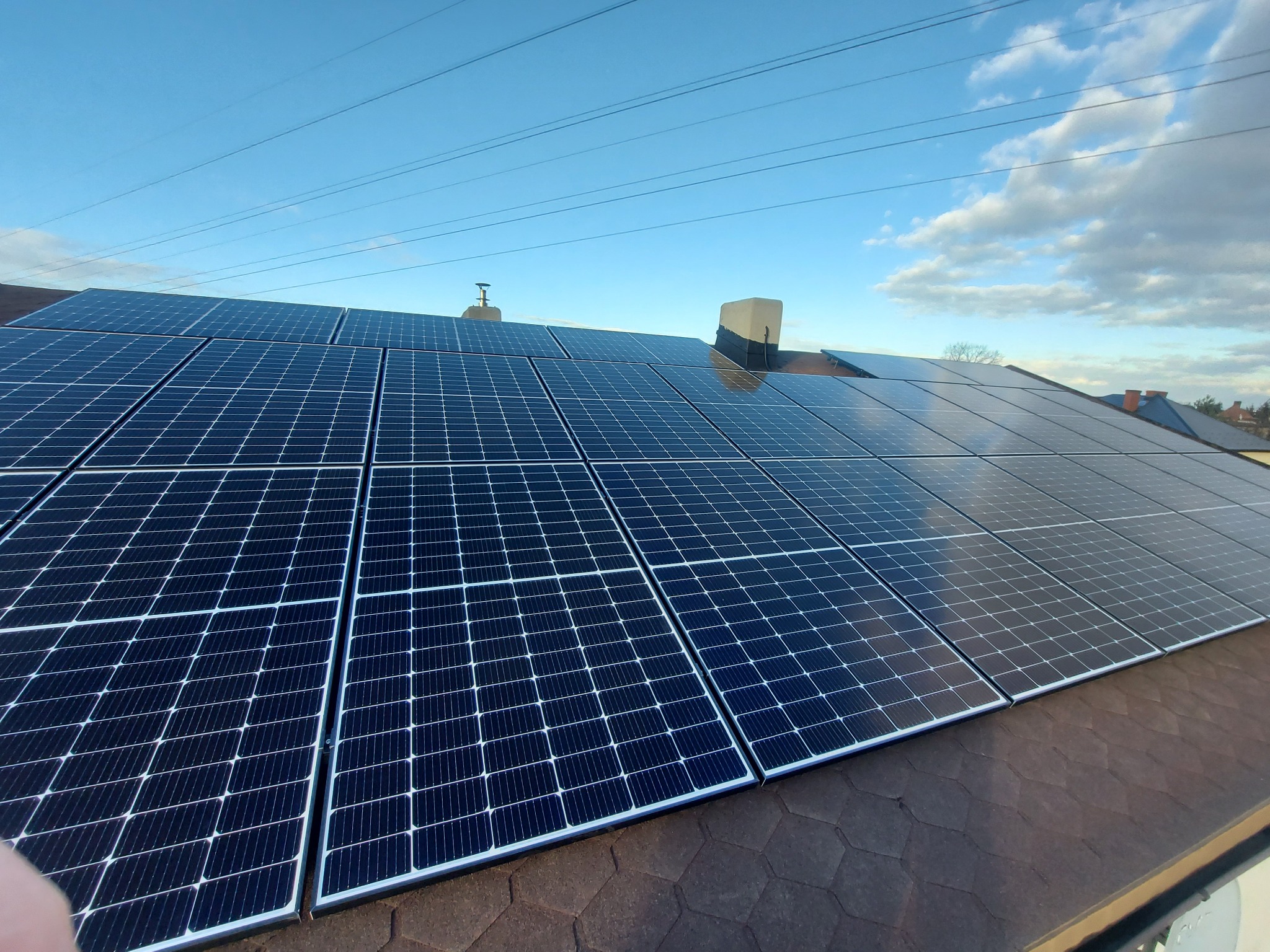 Panel słoneczny na dachu pokrytym gontem bitumicznym, błękitne niebo z lekkimi chmurami w tle.