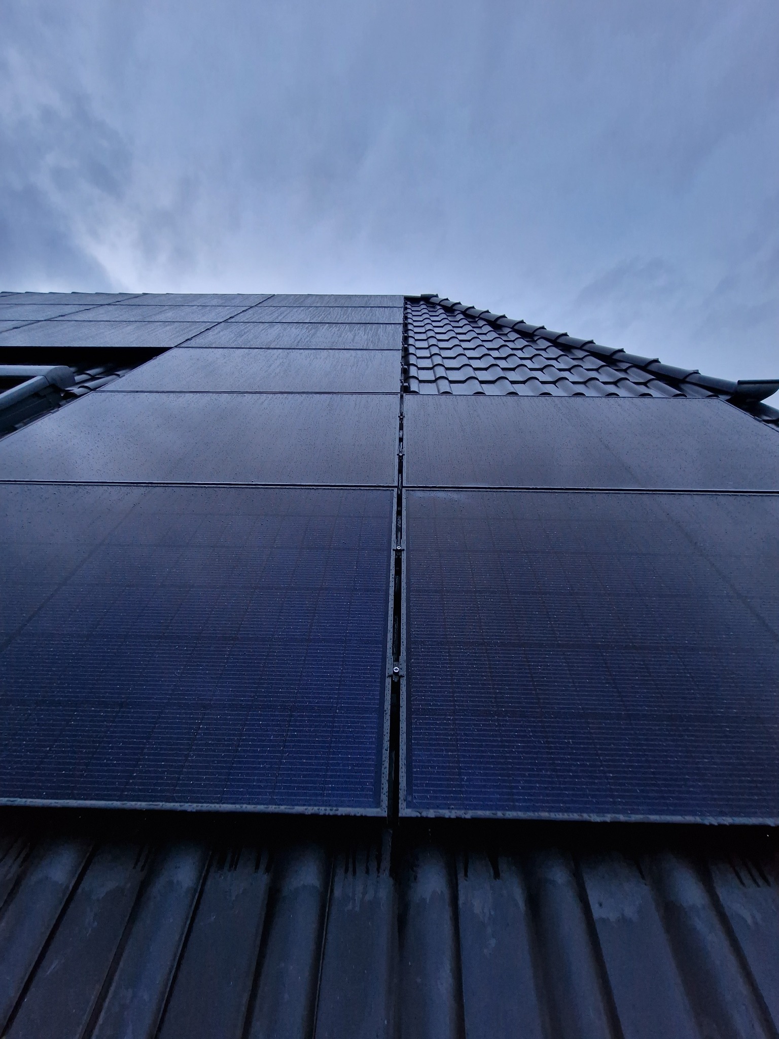 Ciemne panele na dachu pokrytym kroplami deszczu, widoczne fragmenty dachówki i pochmurne niebo w tle.