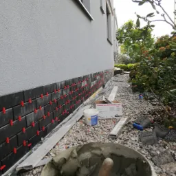 Montaż elewacyjnych płytek klinkierowych na cokole budynku z użyciem czerwonych krzyżyków dystansowych; widoczny pojemnik z zaprawą klejową, puszka z gruntem i opakowanie płytek firmy Roben.