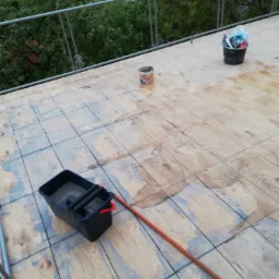 Drewniana podłoga tarasu w trakcie czyszczenia z widocznym wiadrem z wodą, mopem i pozostałościami po farbie oraz wiaderkiem z narzędziami w tle.