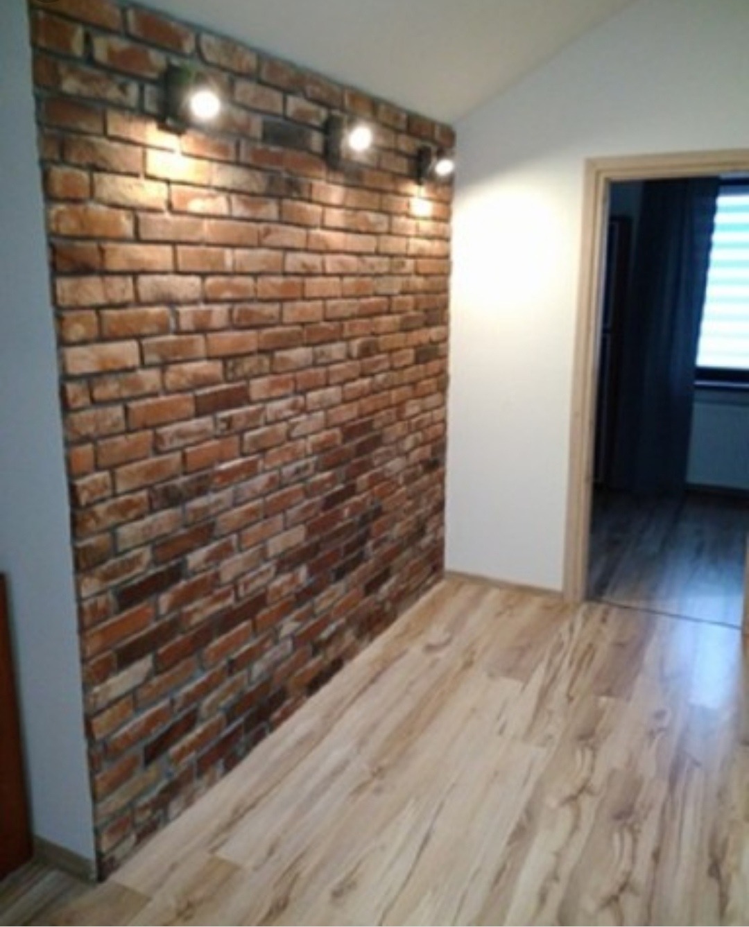 Wnętrze z ścianą wyłożoną cegłą klinkierową, oświetloną trzema reflektorami, oraz podłogą z paneli laminowanych w odcieniach drewna, widok na otwarte drzwi do innego pomieszczenia.