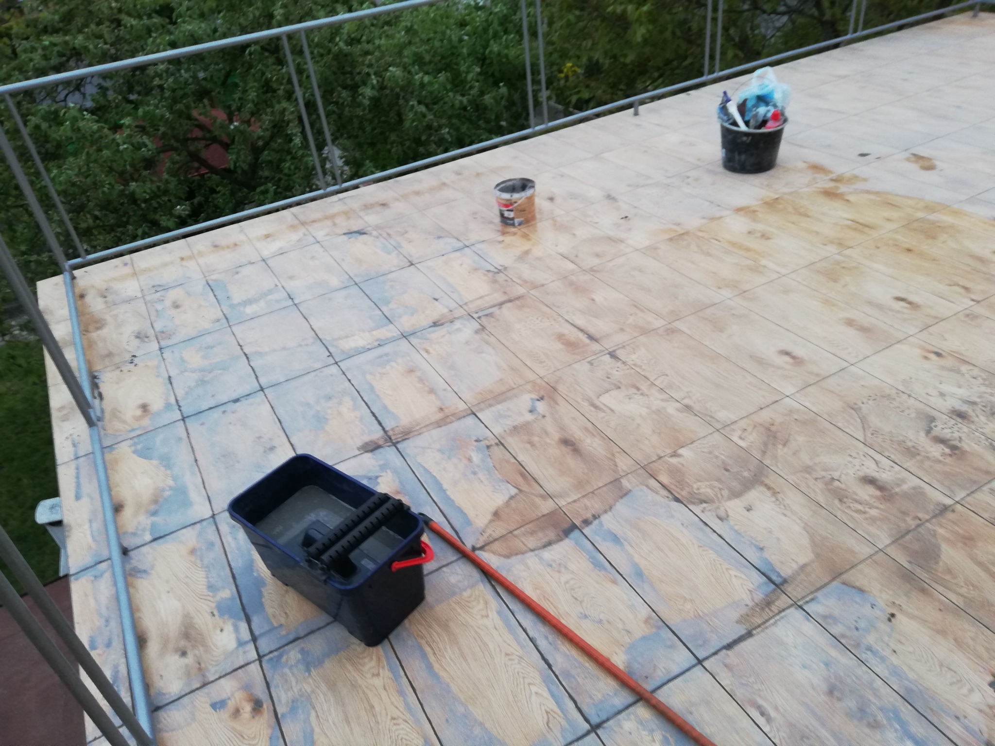 Drewniana podłoga tarasu w trakcie czyszczenia z widocznym wiadrem z wodą, mopem i pozostałościami po farbie oraz wiaderkiem z narzędziami w tle.