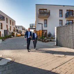 Dwoje agentów nieruchomości w garniturach idzie chodnikiem na tle nowoczesnych budynków mieszkalnych z balkonami i metalowymi ogrodzeniami.