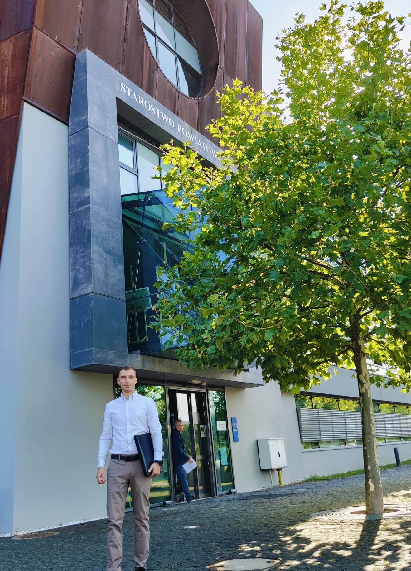 Mężczyzna w koszuli z teczką przed budynkiem Starostwa Powiatowego z nowoczesną architekturą i drzewem liściastym.